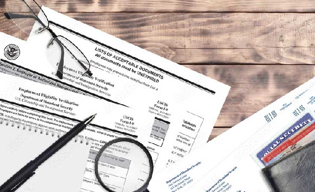 Documentos relacionados à verificação de elegibilidade para emprego, incluindo formulários e materiais de identificação, em uma superfície de madeira.
 Documentos relacionados à verificação de elegibilidade para emprego, incluindo formulários e materiais de identificação, em uma superfície de madeira.