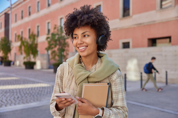 Jovem mulher sorridente usando fones de ouvido, segurando um celular e um caderno, em um ambiente urbano
